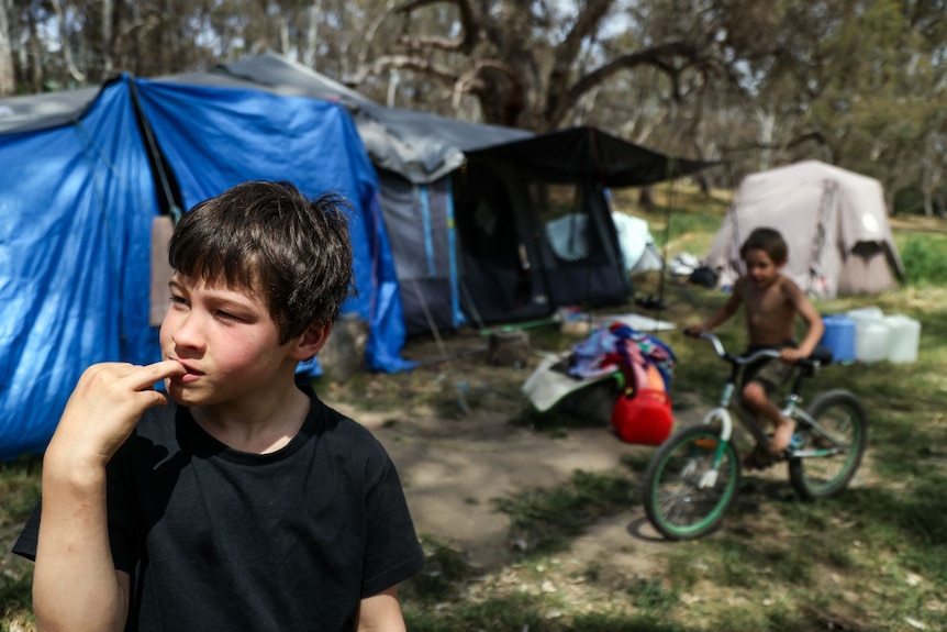 A boy biting a fingernail outside a tent, while his little brother rides a bike in the background.