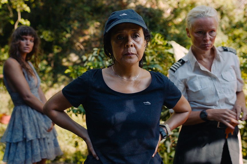 A young woman in a fancy dress, an older woman in cap and tshirt and a middle aged woman in uniform stand in a forest
