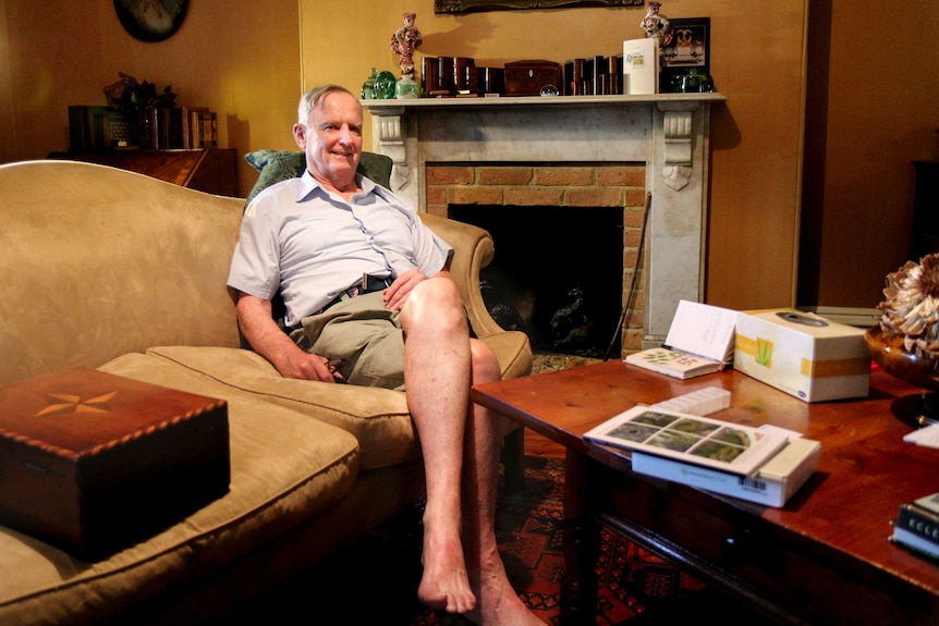 James Guest, wearing shorts and a shirt, sits on a plush couch with a fireplace in the background of an ornate sitting room.