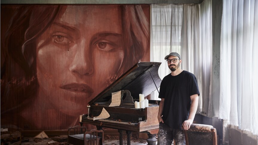 A man leans against a chair in a room filled with autumn leaves and a broken-down grand piano, with a mural of a woman's face.