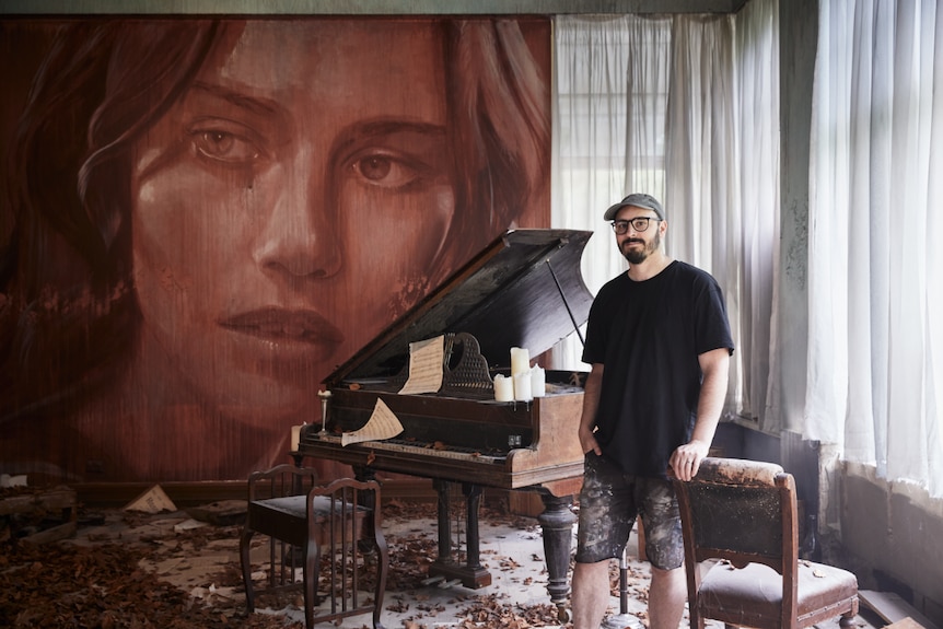 A man leans against a chair in a room filled with autumn leaves and a broken-down grand piano, with a mural of a woman's face.