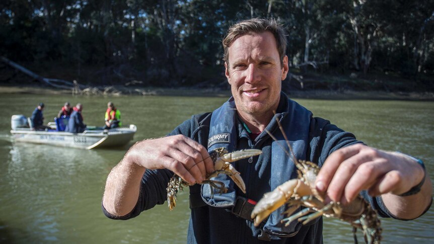 Nick Whiterod holds two Murray crayfish on the banks of the river, while four men sit in a tin boat on the water behind him.