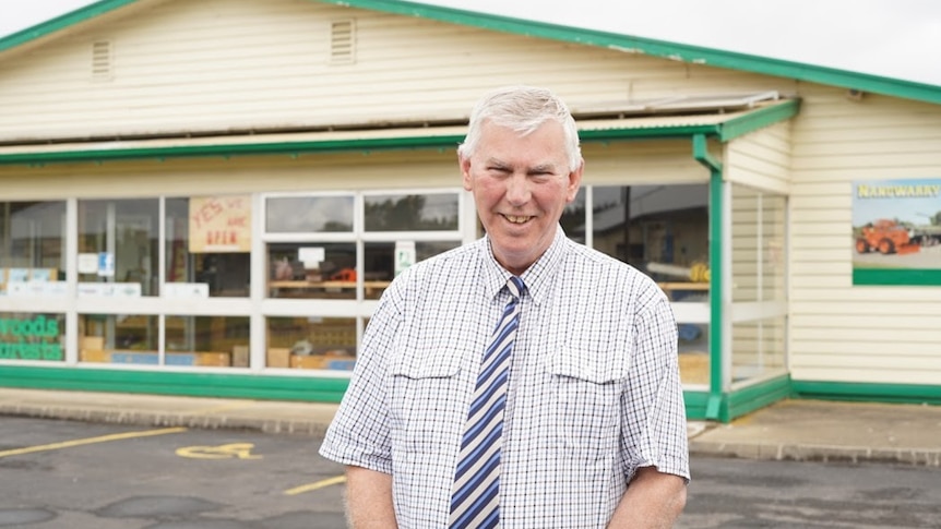 A man in a short-sleeved white and blue buttoned shirt smiles in a carpark.