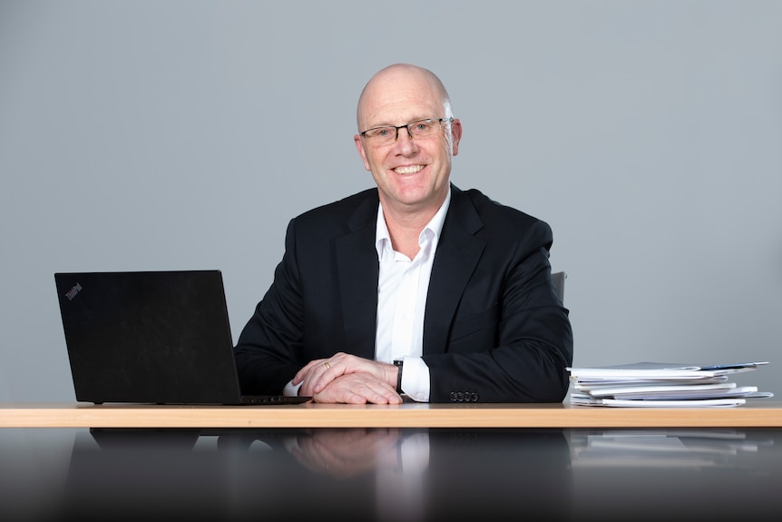 a man wearing glasses sitting at a desk behind a laptop and a stack of papers smiling at the camera