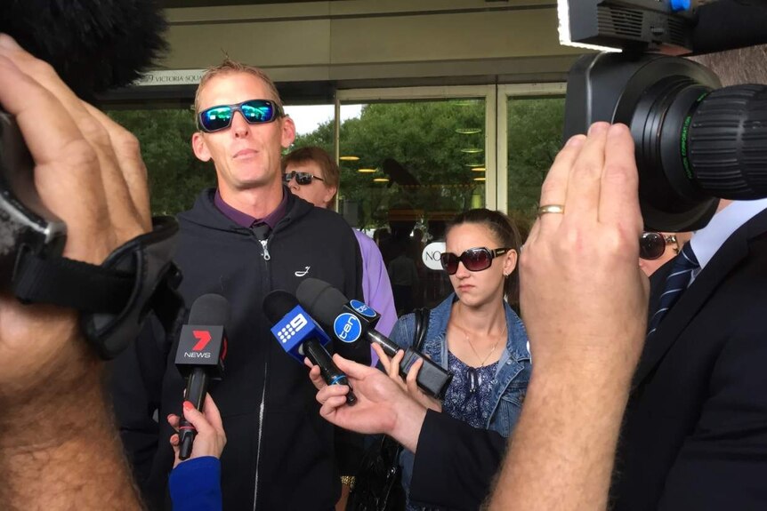 A man in sunglasses is surrounded by media cameras and microphones.