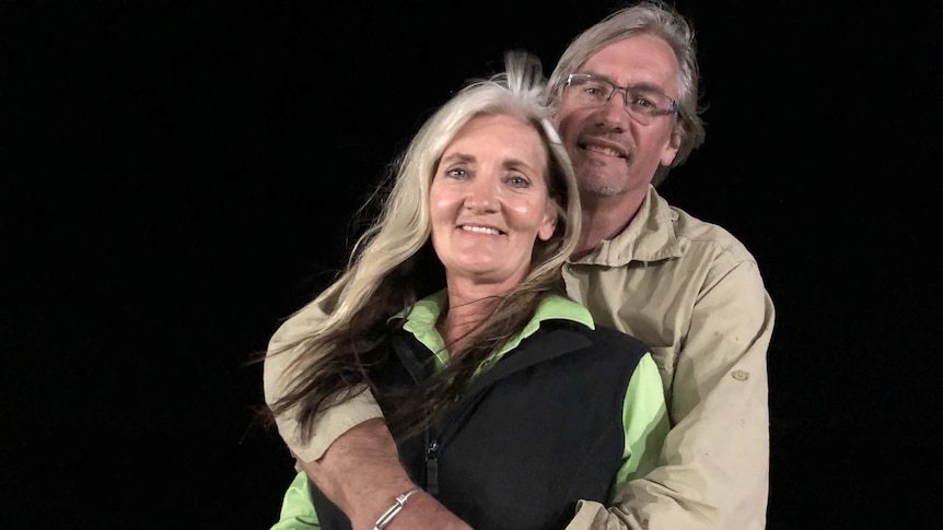 A woman with long white hair smiles and a man with glasses, white hair and a khaki shirt smiles with his arms around her.