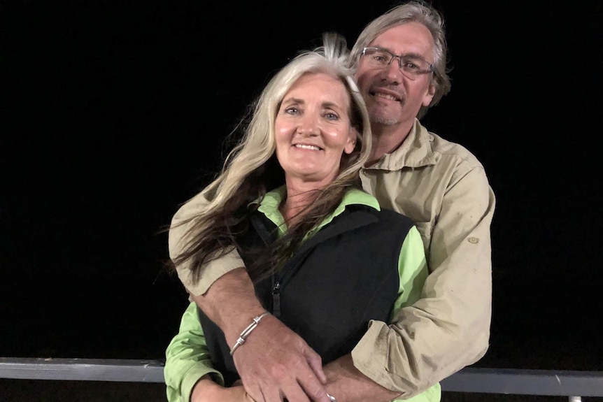 A woman with long white hair smiles and a man with glasses, white hair and a khaki shirt smiles with his arms around her.