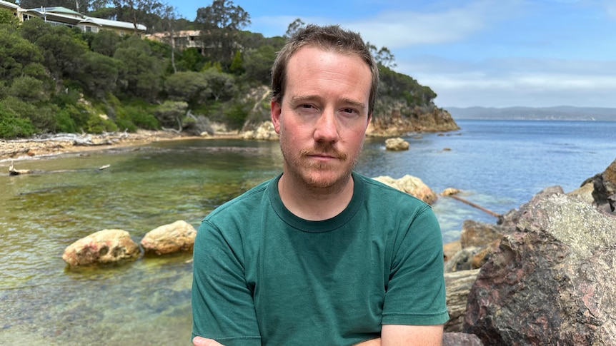 A man in a green shirt looks at a camera seriously with a nice bay in the background.