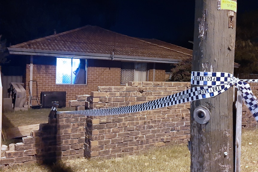 A house at night behind a brick wall with police tape around a power pole.