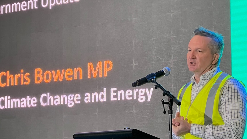 A man dressed in high-vis with short, dark hair – Energy Minister Chris Bowen – speaks at a lectern next to a large screen.