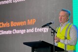 A man dressed in high-vis with short, dark hair – Energy Minister Chris Bowen – speaks at a lectern next to a large screen.