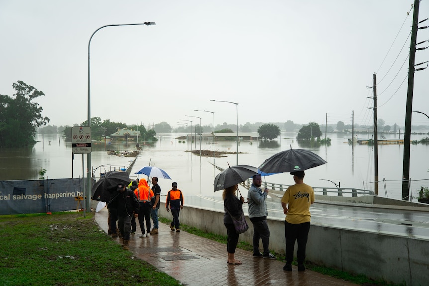 People gather under umbrellas at the entrance to a bridge that is under water.