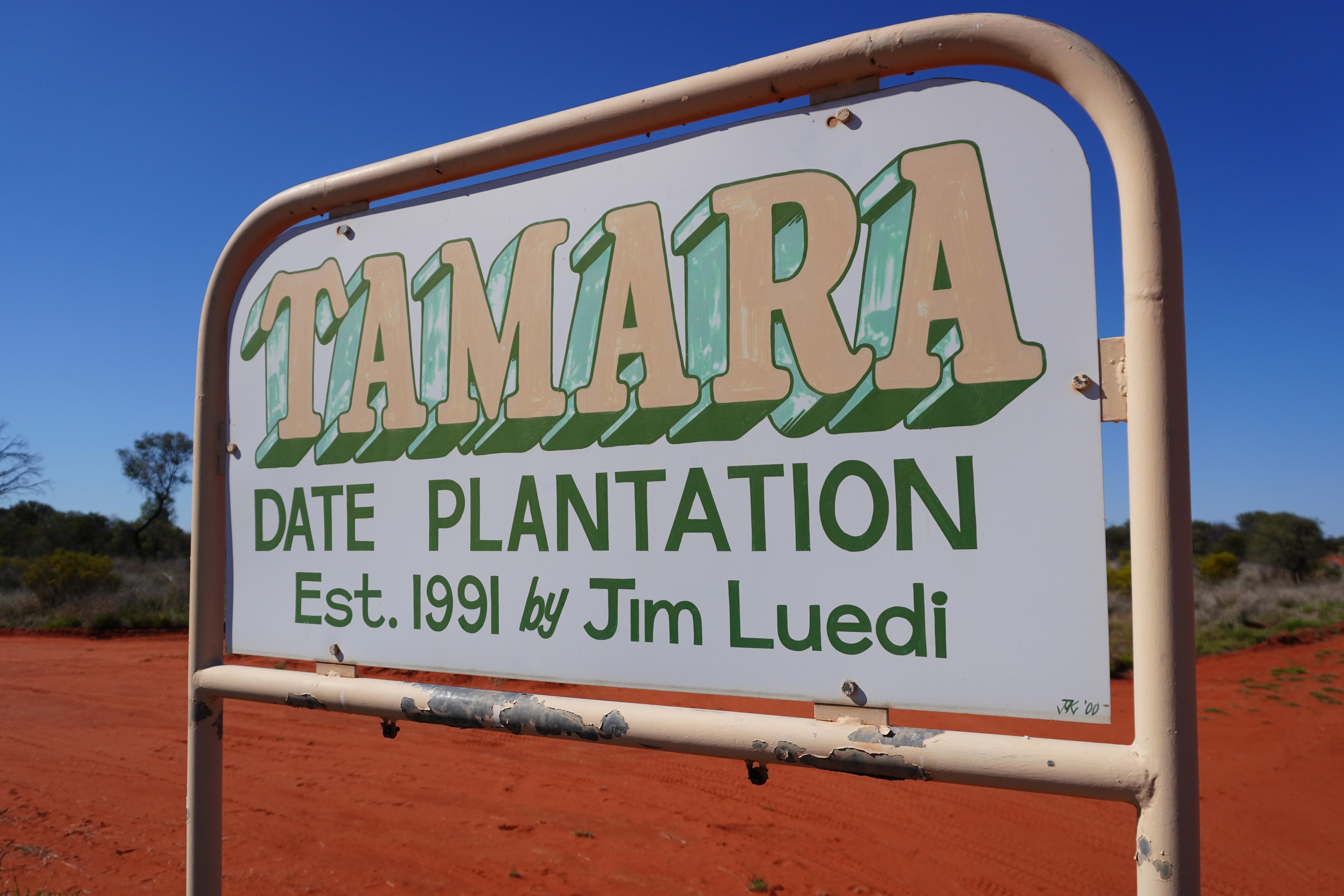 A sign for the Tamara Date Plantation, with a red dirt, desert road in the background.