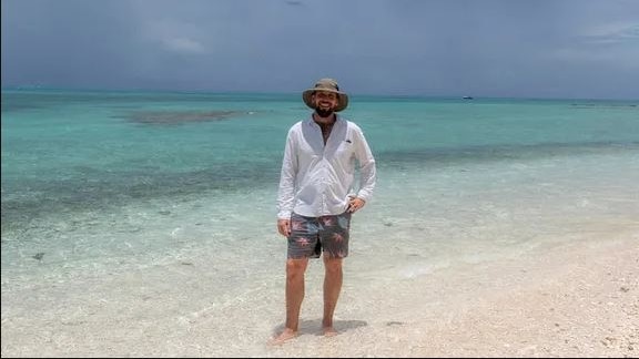A man in hat, boardies, and a long-sleeved shirt stands, smiling, in clear shallow waters at the beach.