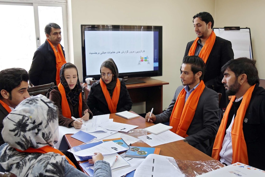 In a small office, a mix of men and women in orange scarves listen to a women in the middle around a kitchen-size table.