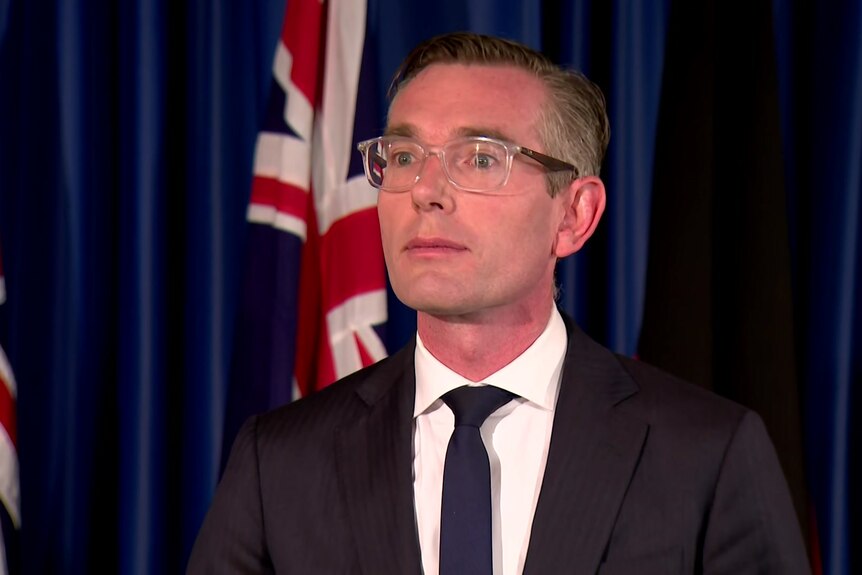 Man with glasses wearing suit and tie stand in front of navy backdrop and two australian flags