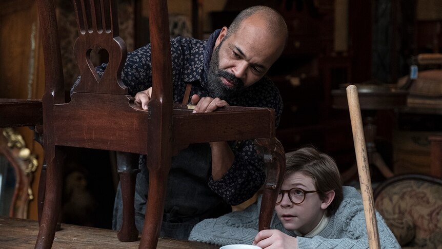 A man with a beard works closely on a wooden chair in a workshop as a young bespectacled boy looks on.