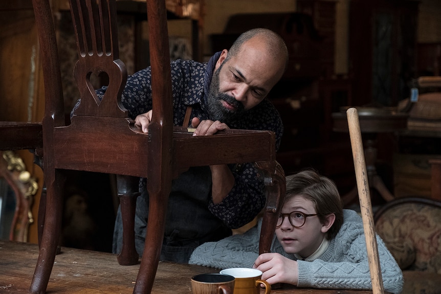 A man with a beard works closely on a wooden chair in a workshop as a young bespectacled boy looks on.