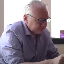 Julian Assange with white hair and glasses in a blue shirt reads documents on a couch