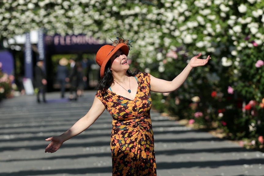 A woman in a patterned orange and black dress smiles in the sunshine under a rose-adorned archway.