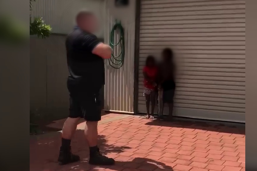 A blurred image showing two children against a garage door, and a man next to then with arms crossed. 