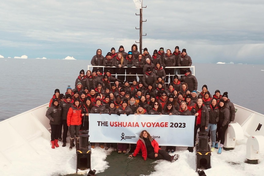 91 female scientists from around the world stand at the bow of the ship holding a sign that says the USHUAIA voyage.