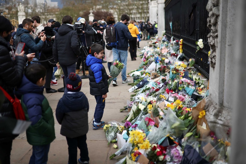 A child wears a blue and black puffer jacket and is seen holding a bouquet of flowers as he walks up to lay them among tributes.