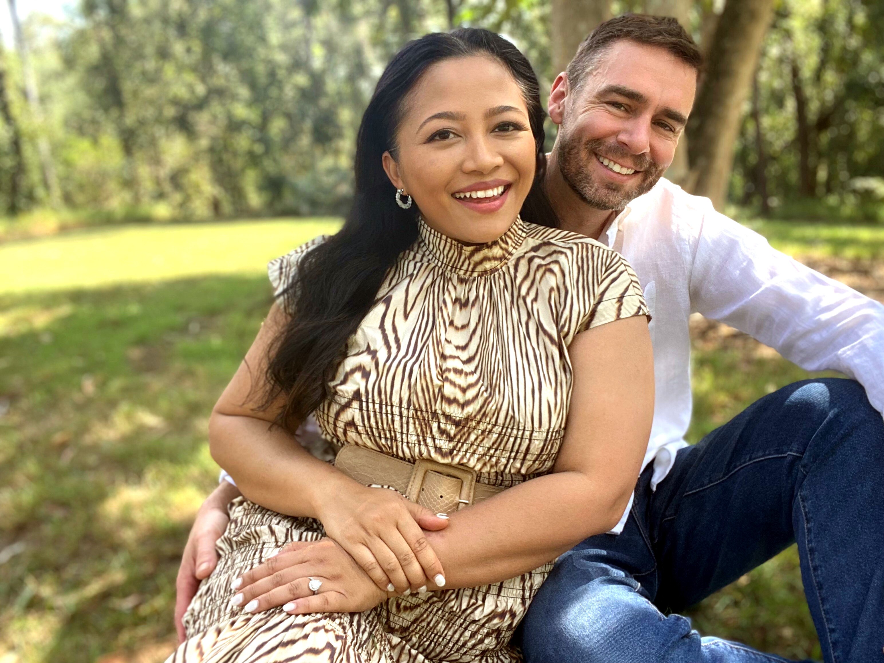 Donna and Greg smile while sitting outside on a sunny day. Donna is wearing an engagement ring.