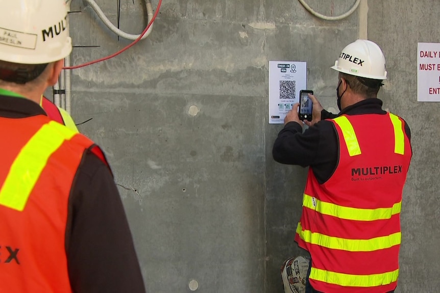A worker in hi-vis vest holds a mobile phone up to a QR code sign at a building site.