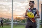 A young Indigenous AFL player smiles as he spins a yellow football in his hands in front of goalposts.