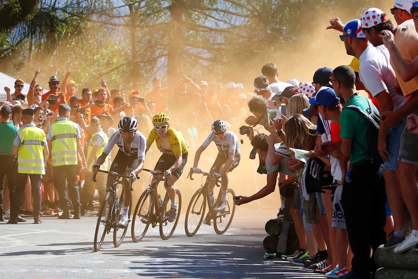 Men ride bikes past cheering spectators behind a haze of orange smoke.