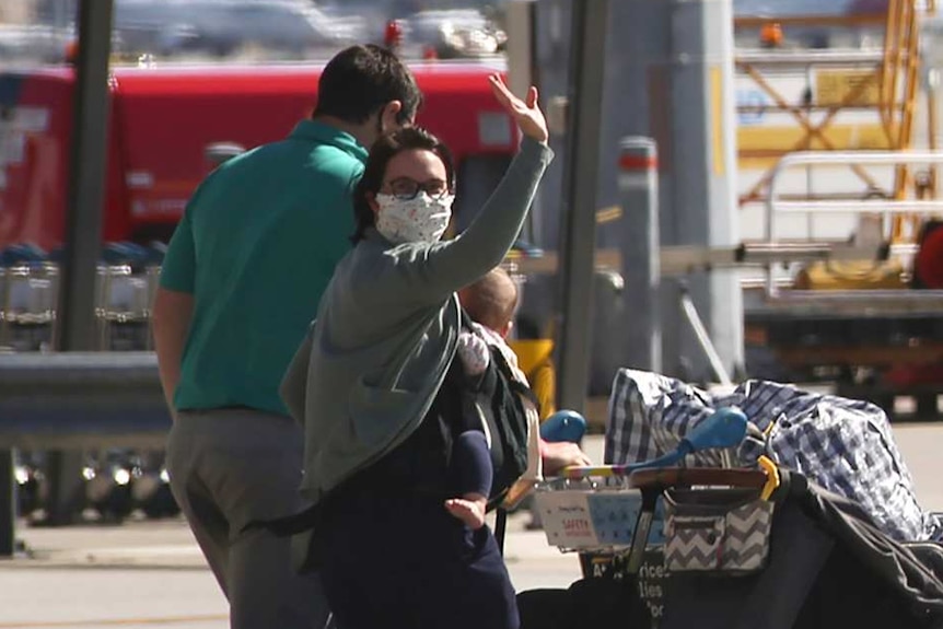 Passengers walk across the tarmac at Perth Airport pushing trolleys and carrying luggage as a woman in a face mask waves.