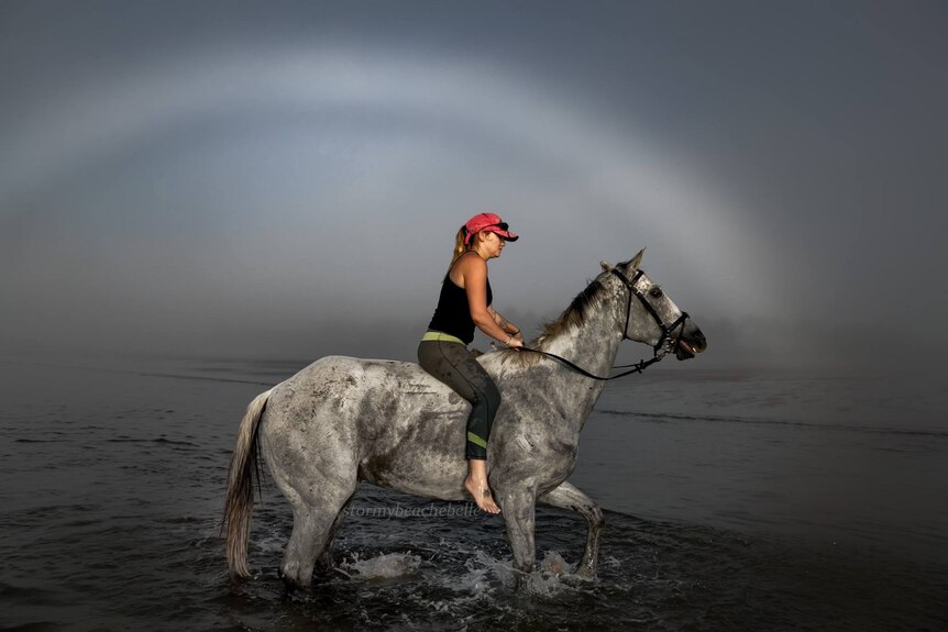 A horse rider framed by a fogbow in the water at the beach.