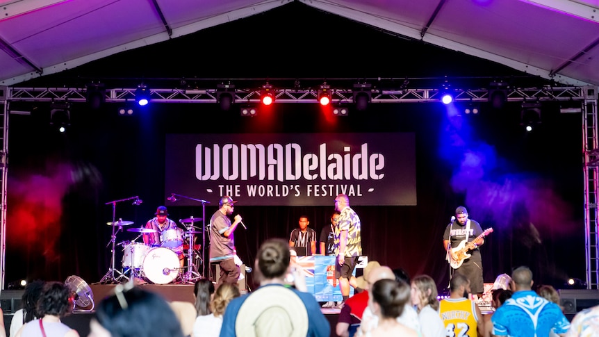 A band performs onstage at WOMADelaide. A crowd is seen in front of the stage.