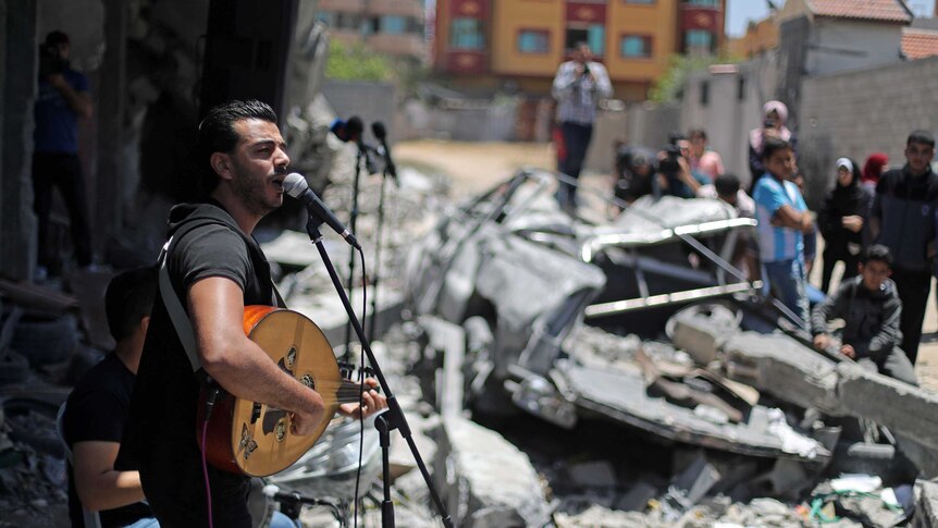 A man sings at a microphone surrounded by rubble from buildings.