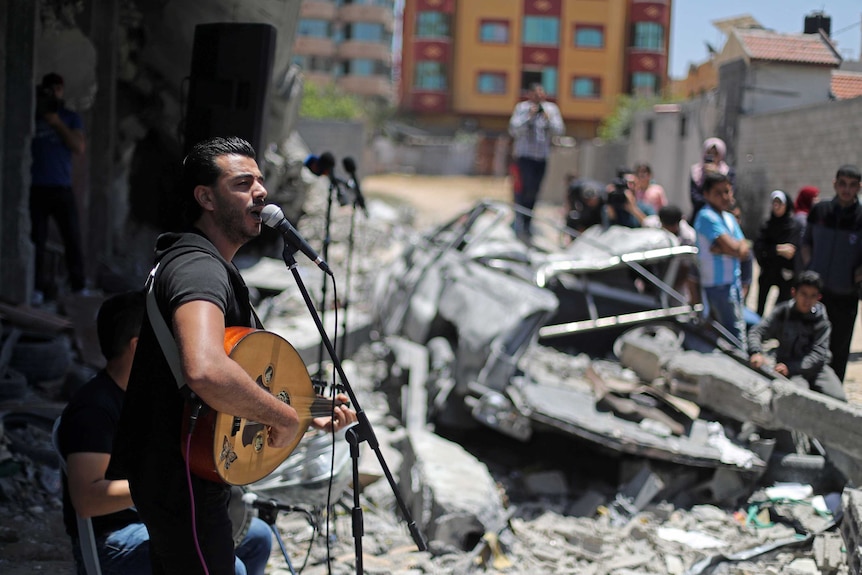 A man sings at a microphone surrounded by rubble from buildings.