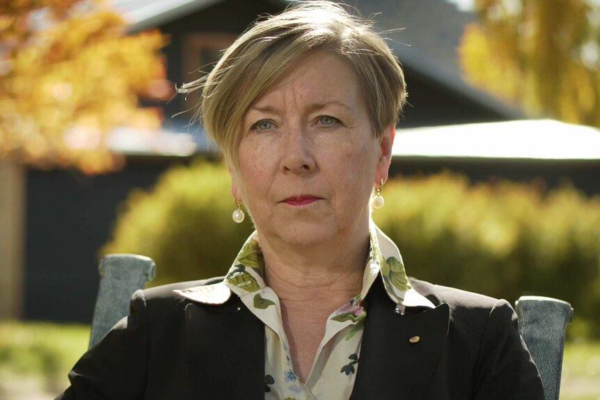 Jane Halton wearing a brown jacket and light green floral shirt, sitting outside with autumnal trees and bushes behind her