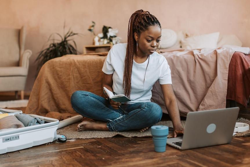 A young woman in white t-shirt and blue jeans sits on the floor next to an open suitcase, typing on a laptop