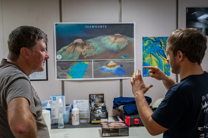 An older man listens to a younger man explain seamounts with posters of the mountains in the background