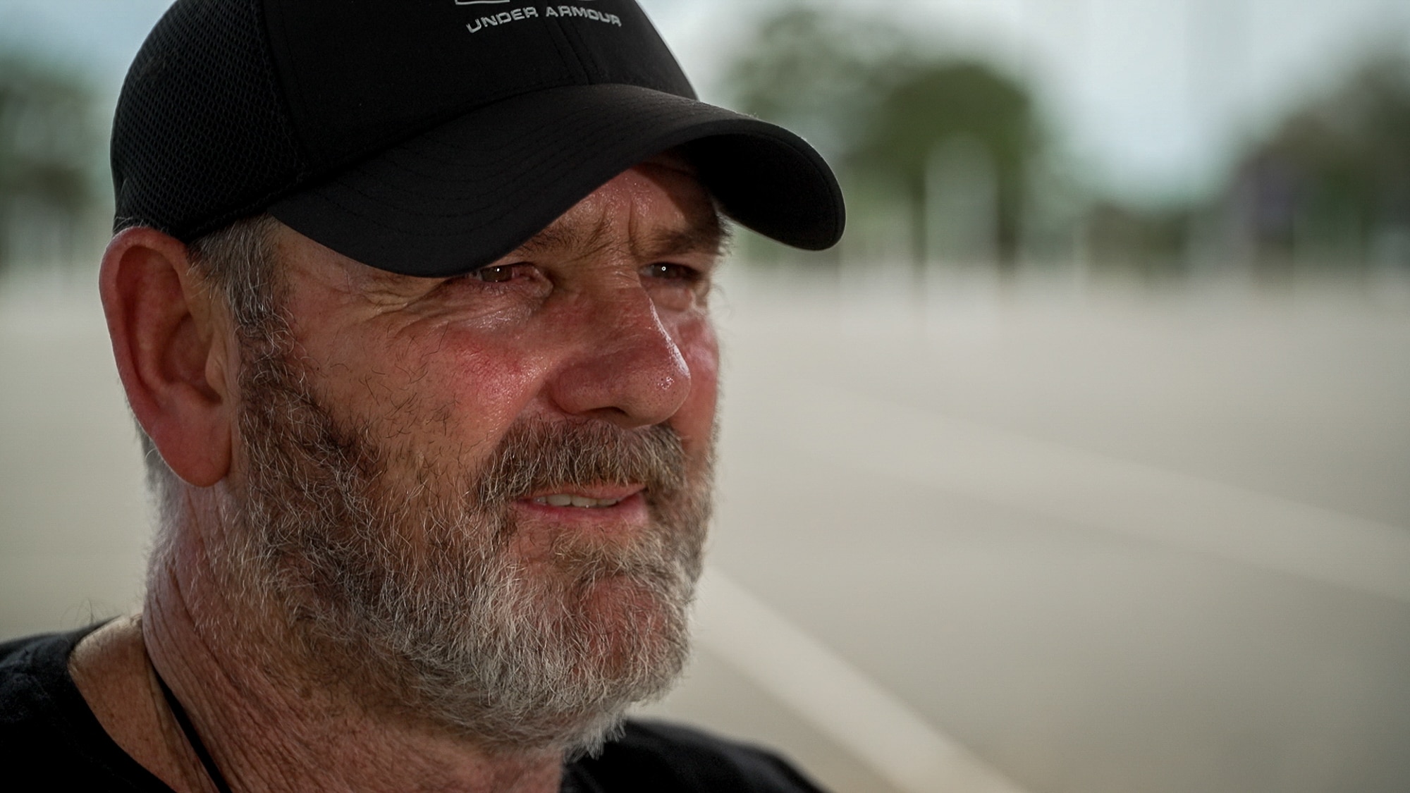 Um close mostra um homem com barba grisalha, usando um boné desbotado, olhando para longe