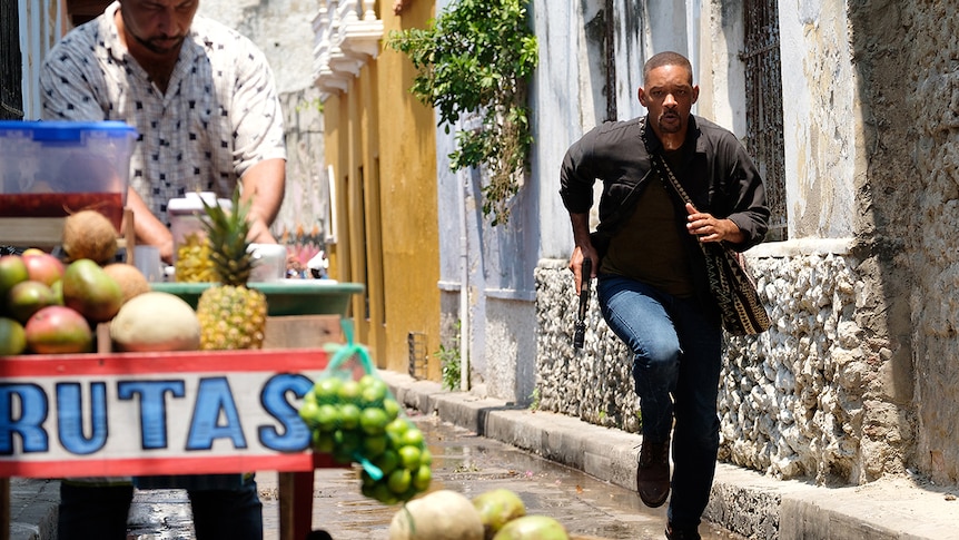 Will Smith runs down narrow street on a sunny day, with pistol in hand and bag over shoulder near a street vendor.