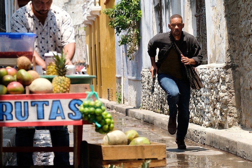 Will Smith runs down narrow street on a sunny day, with pistol in hand and bag over shoulder near a street vendor.