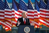 A man wearing a dark suit with a red tie holds his hands in the air while standing at a podium with US flags behind him.