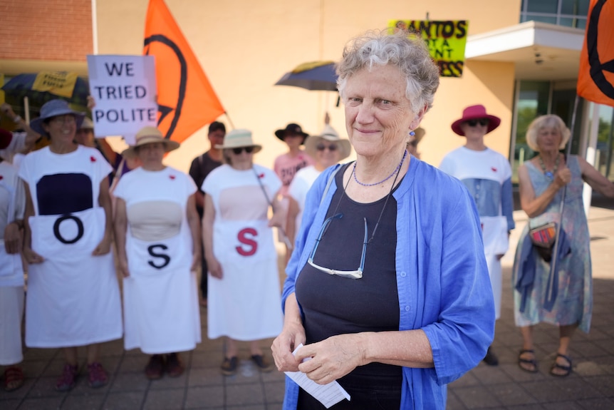 A woman with white and grey hair stands in front of protestors, one holds a sign that reads 'we tried polite'