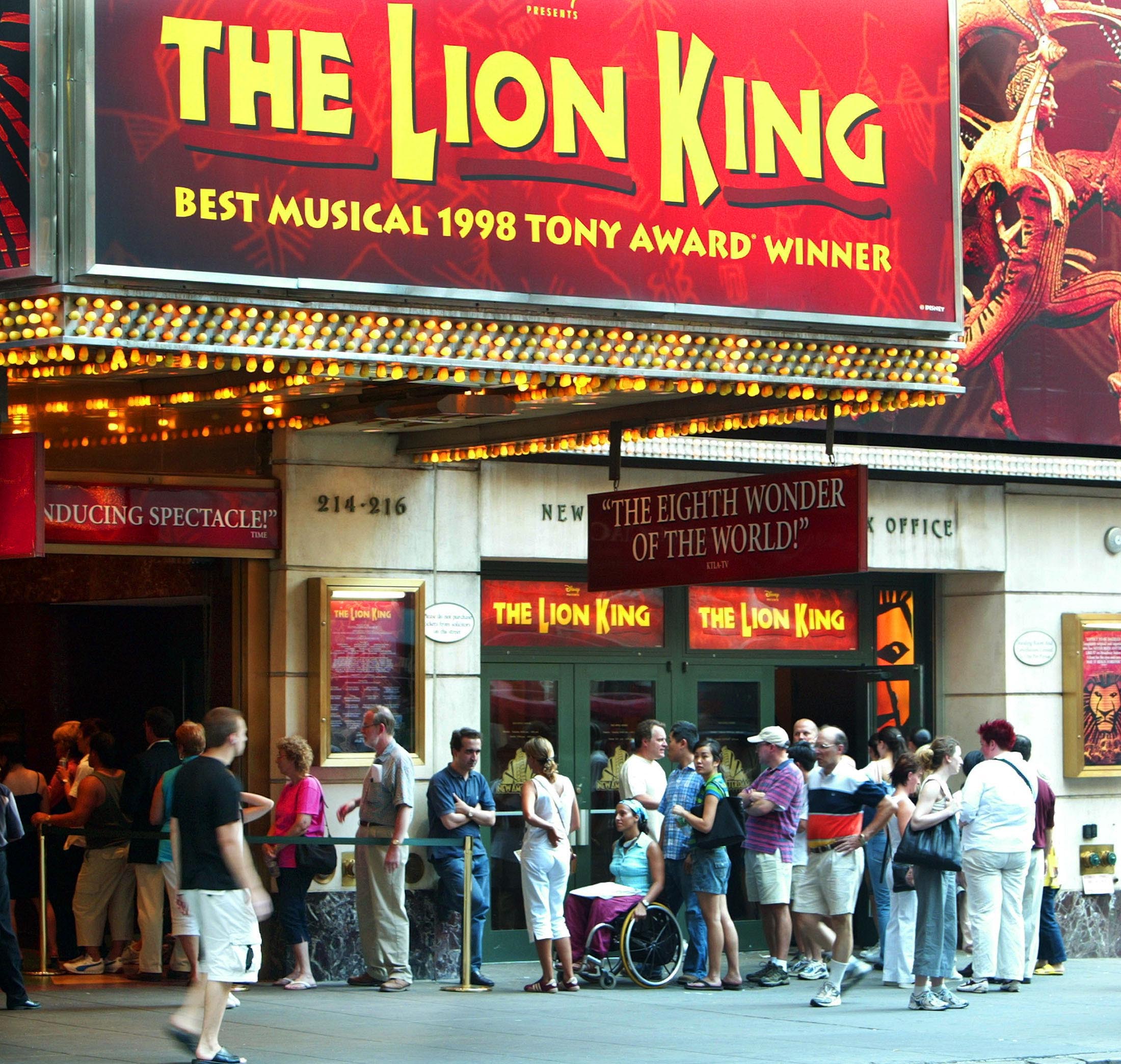 Várias pessoas fazem fila em frente à bilheteria da Broadway com uma placa gigante que diz O Rei Leão acima dela.