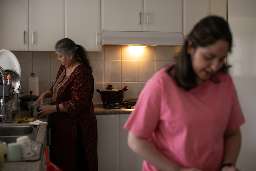 An older and younger woman, one blurred, in the kitchen, pot on stove. Younger wears pink tee, older, Indian clothes.