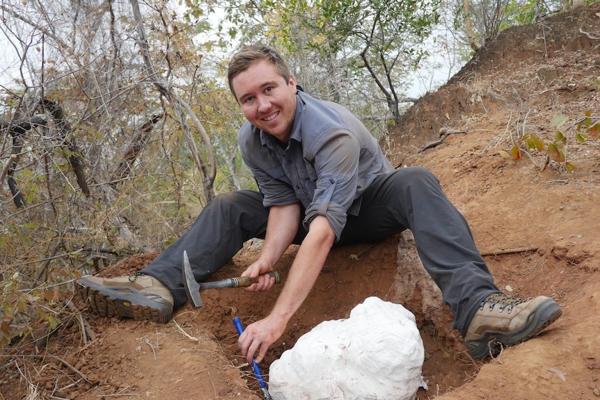 A man is seen with a hammer and tool, excavating a part of a skeleton that is sitting in a hole on a mound of light brown dirt. 