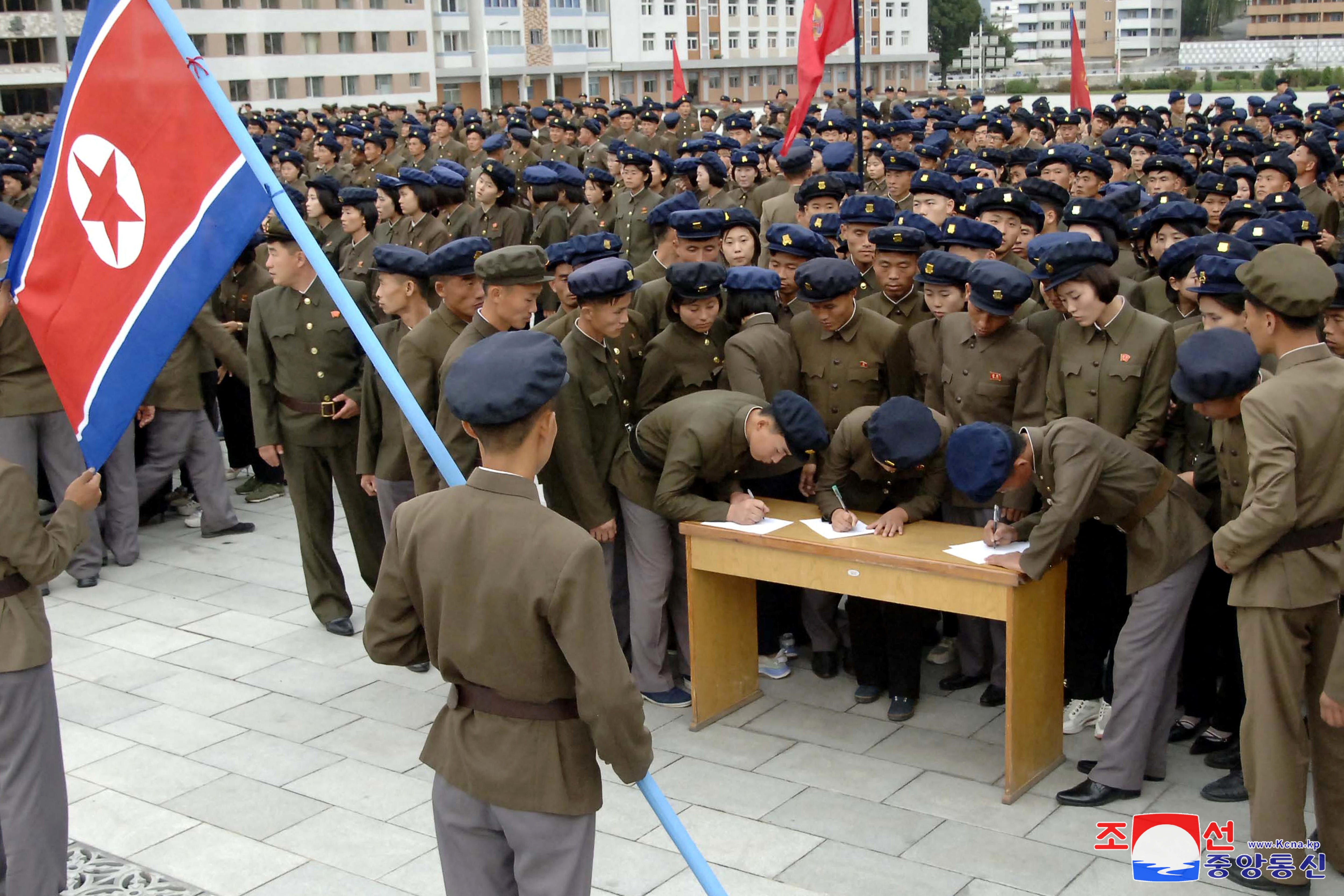 Hundreds of people in army uniforms stand in an outside square congregating toward a table where people are signing forms