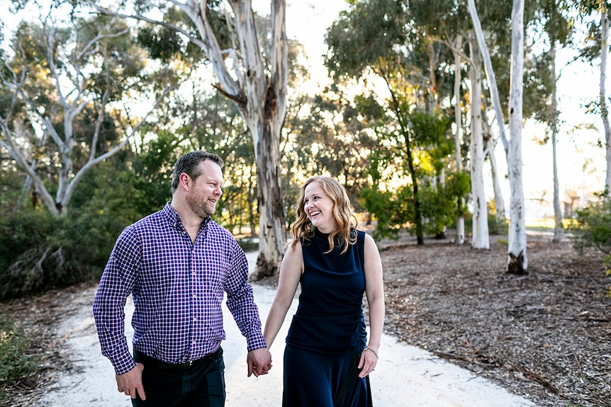 A man and woman, holding hands and smiling at with gum trees behind them. The couple wants an unplugged wedding.