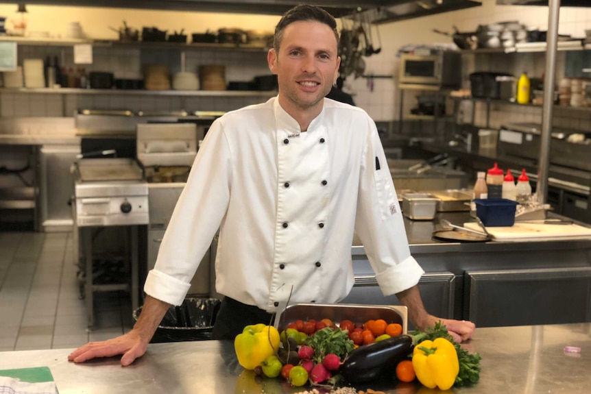 A man wearing a white chef's shirt stands in front of a bowl of vegetables